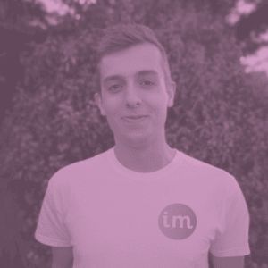 Portrait of Joe Lamyman, smiling with his brown hair swept to the side. The photo captures Joe from the torso up. Joe is wearing a white t-shirt with a rounded logo, feature the letters 'im' on the left breast. Joe is stood in front of a large bush, covering the background. A pink and purple filter has been applied to the photo.