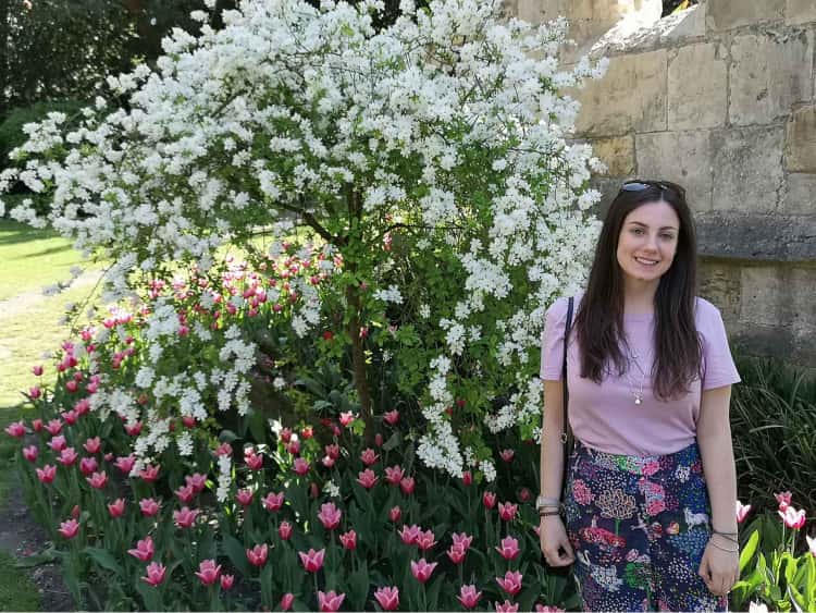 Naomi stood in front of some beautiful pink spring flowers, with a bush flowering with bright white flowers behind her.