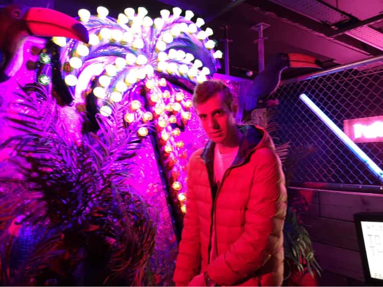 Myself in a bright orange jacket, in front of a tropical lit up background at Junkyard Golf in Leeds. I look very focussed or determined. Either way, the look did not help my golf performance.