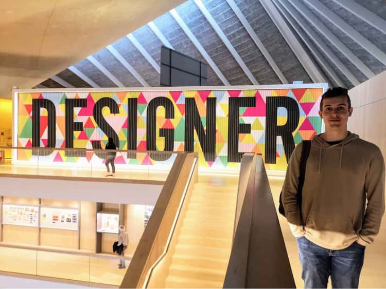 Myself stood on the top floor of the Design Museum in London, with a large display in the background (the height of the walls) displaying the word DESIGNER, with a geometric pattern surrounding it.