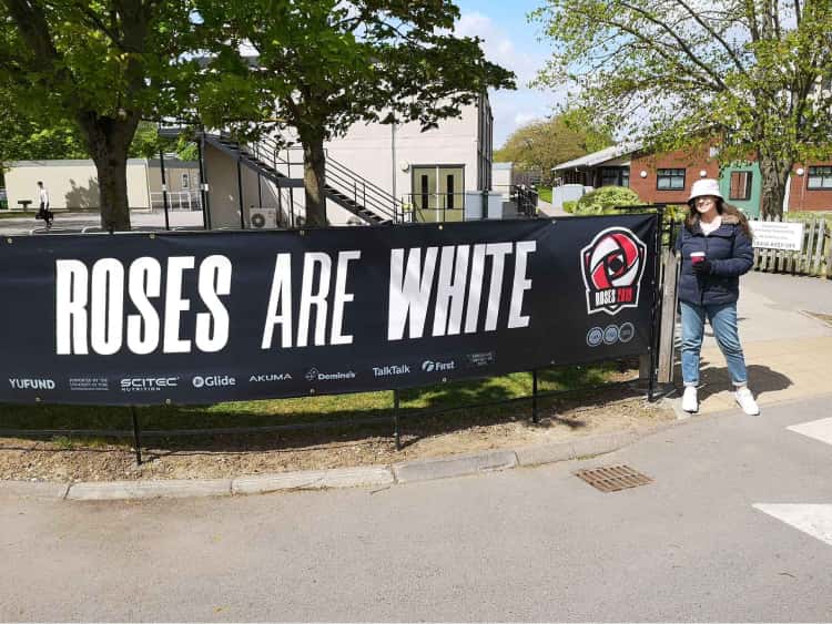 Naomi in her winters coat stood next to a banner that says, Roses are white, on the University of York campus.